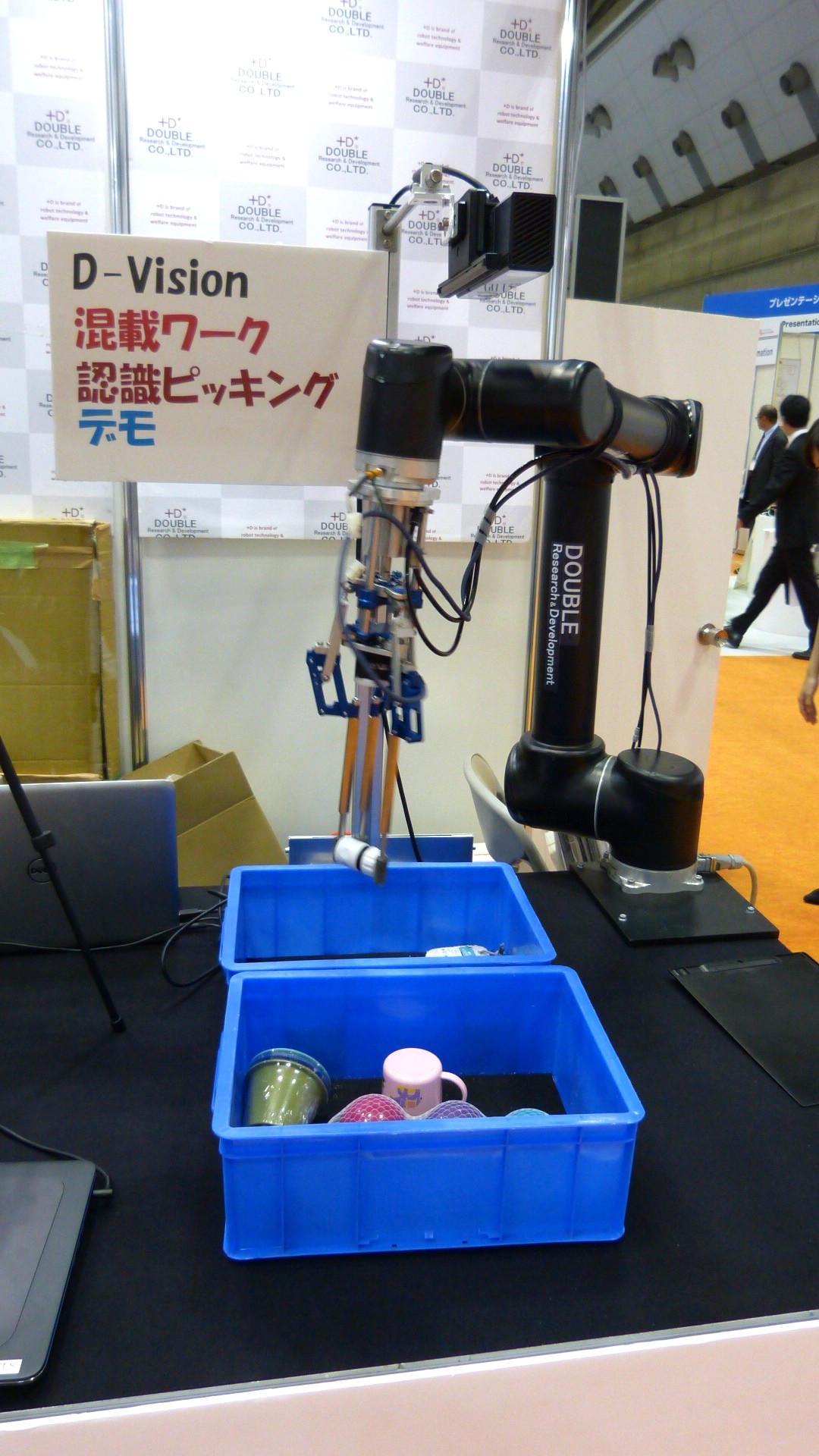 a robot arm sorts coffee mugs from one bin into another near a sign that reads D-Vision